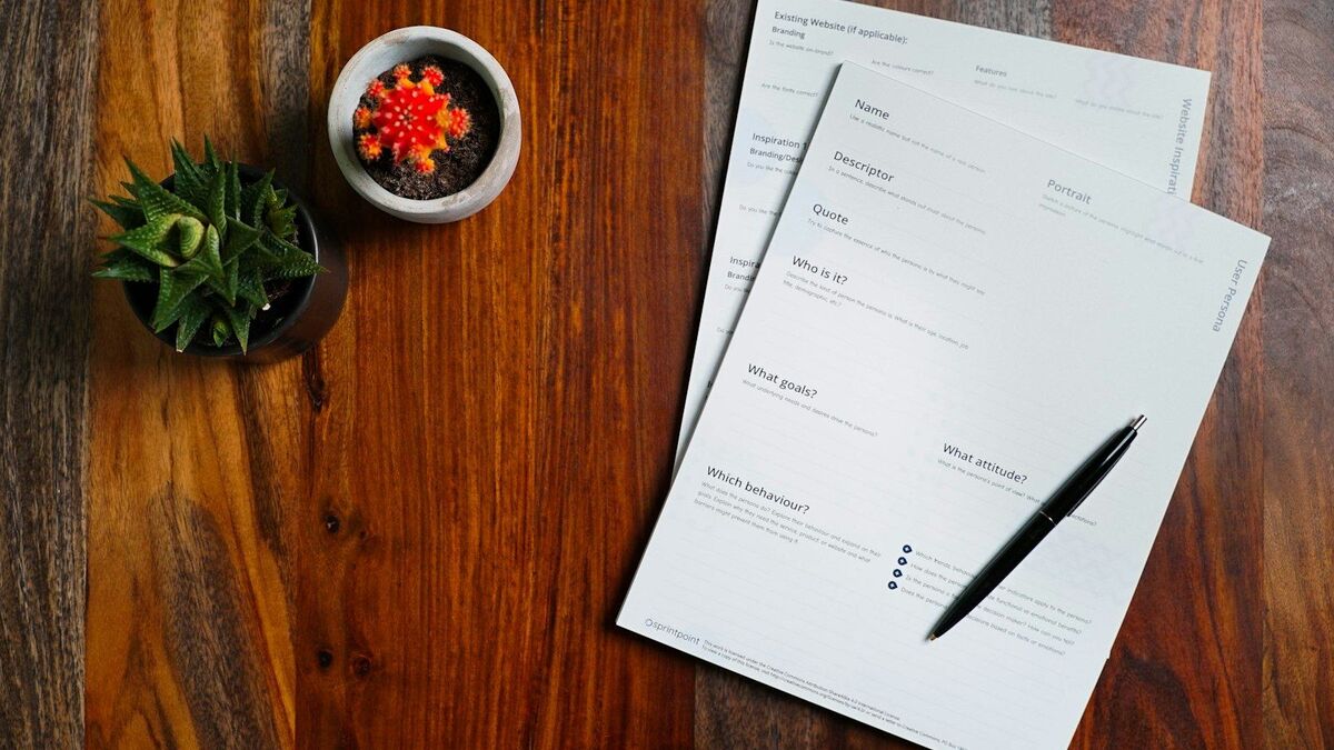 An overhead view of a timber desk with printed forms, a pen and two small potted plants.